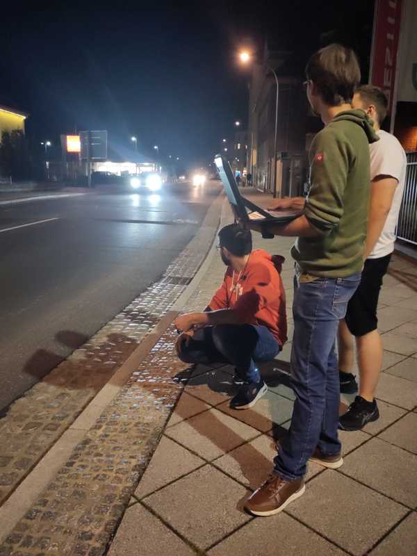Ein Team steht nachts draußen an der Straße und testet ihr System. Man sieht rechts im Vordergrund die Teammitglieder mit einem Laptop stehen. Links ist die Straße, auf der man in einiger Entfernung Autos mit grellen Scheinwerfern heranfahren sieht