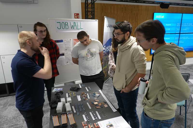 Marco vom Orga-Team erklärt einigen Teilnehmenden die Sensoren und Mikrocontroller. In der Mitte sieht man einen Tisch, auf dem alle Sensoren, Kabel und weitere Materialien ausliegen. Die Teilnehmenden stehen um den Tisch herum verteilt.