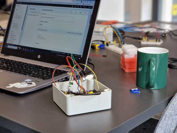 Ein Sensor mit einigen Kabeln steht auf einem Tisch, dahinter ein aufgeklappter Laptop und eine Kaffeetasse. Im Hintergrund sieht man weitere Sensoren, Mikrocontroller und Kabel