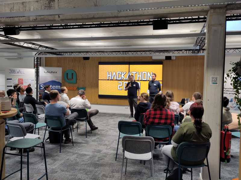 Manuel und Max vom Vorstand des Vereins eröffnen die Veranstaltung. Im Hintergrund sieht man das Logo des Hackathons und im Vordergrund zahlreiche Leute auf Stühlen sitzend.