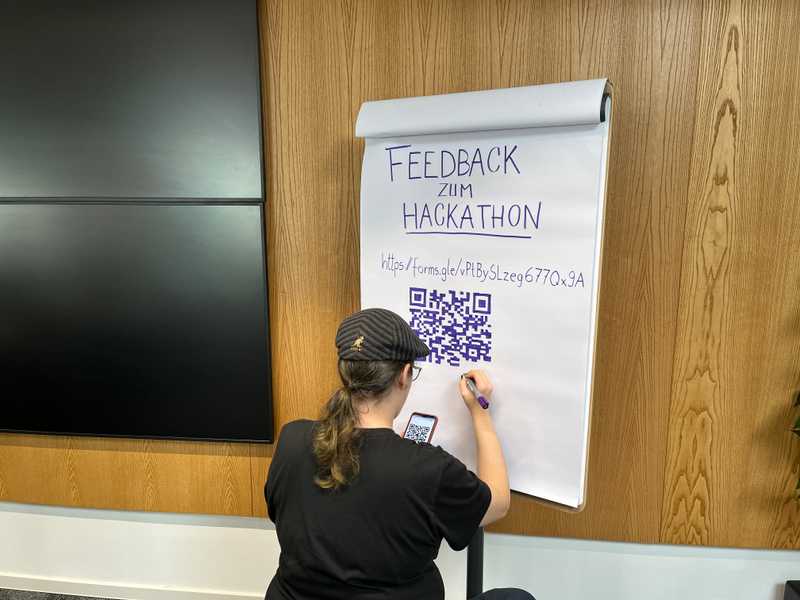 Marlene vom Orga-Team hockt vor einem Papier-Flipchart. Darauf hat sie einen Aufruf zum Abgeben von Feedback gemalt. Nun malt sie einen QR-Code per Hand auf das Papier. Der Link ist "https://forms.gle/vPtBySLzeg677Qx9A"